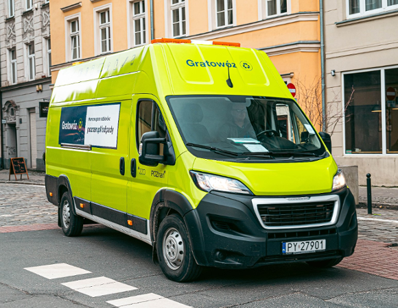Dostawczy samochód marki Peugeot w jaskrawożółtym, seledynowym kolorze, jadący po brukowanej ulicy w mieście. Na dachu samochodu pomarańczowe lampy i duży, zielony napis "Gratowóz". Po bokach reklama i adres strony, w tym: "poznan.pl/odpady".