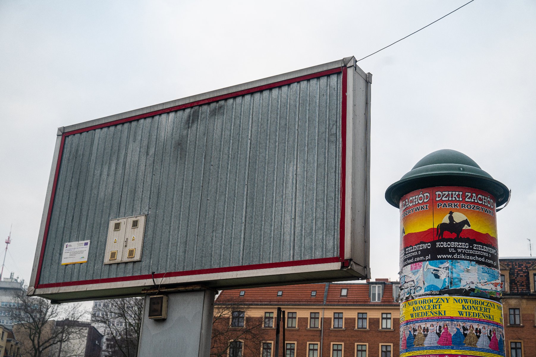 Duży, pusty bilbord z blachy falistej w czerwonej ramie. Po prawej stronie stoi zielony słup ogłoszeniowy z kolorowymi plakatami.