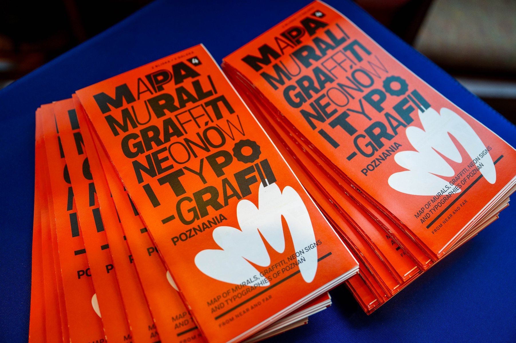 Dwa stosy pomarańczowych ulotek lub broszur rozłożone na niebieskim podłożu. Na okładkach widnieje duży, czarny napis w języku polskim: »MAPA MURALI, GRAFFITI, NEONÓW I TYPOGRAFII POZNANIA«. Poniżej znajduje się biała grafika przypominająca dłoń lub kwiat oraz mniejszy napis w języku angielskim.