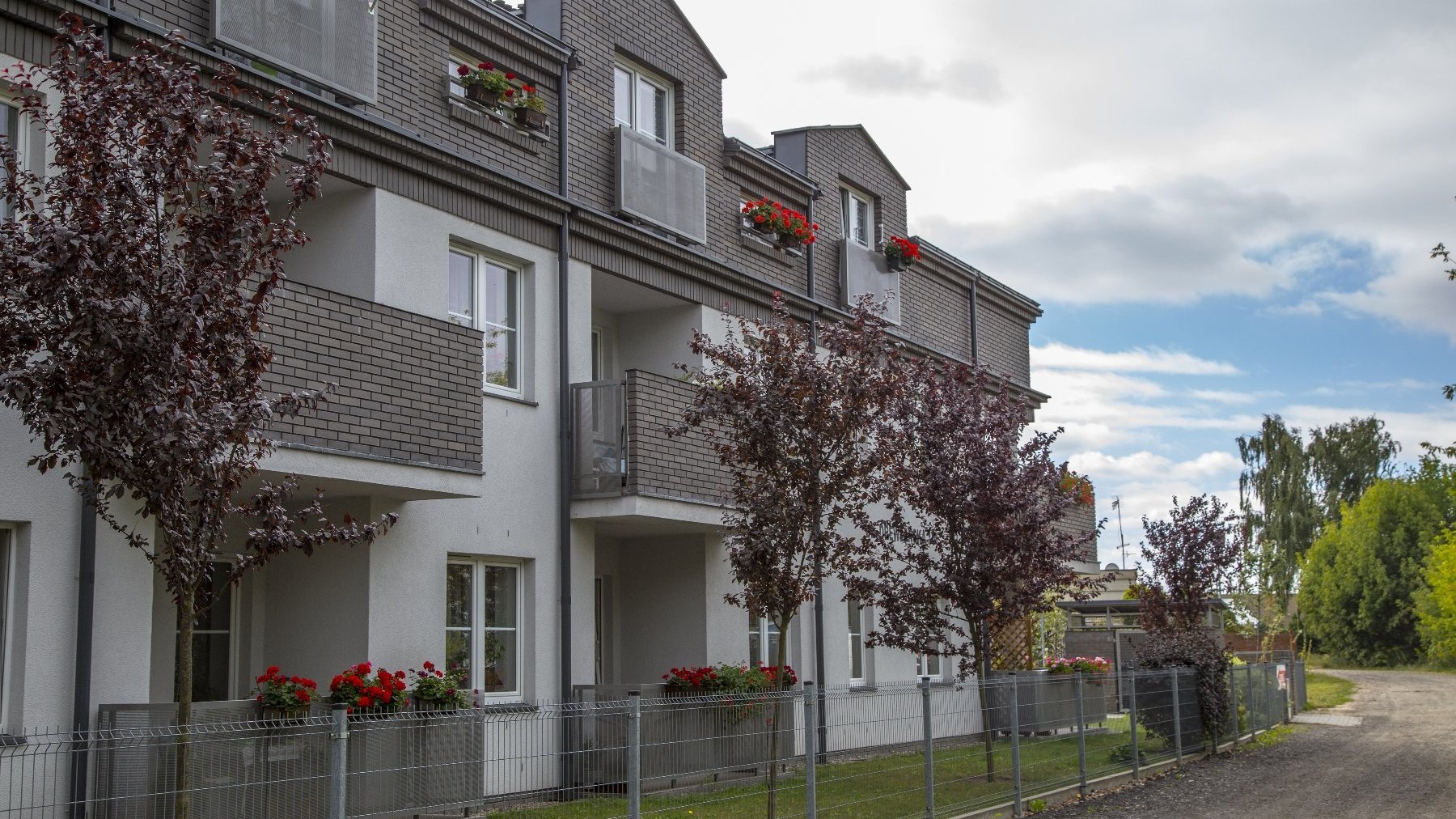 Dwupiętrowy, nowoczesny budynek mieszkalny o białej elewacji z szarymi, ceglanymi wstawkami na piętrach. Przed budynkiem rosną małe drzewa o bordowych liściach. Na balkonach i pod oknami widać doniczki z czerwonymi kwiatami. Na pierwszym planie niski metalowy płot.