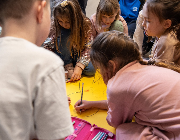 Grupa dzieci siedzi na podłodze i rysuje na dużym żółtym arkuszu papieru. Na pierwszym planie piórnik z kredkami i ołówkami. Dzieci pochylone nad pracą, skupione na wspólnym rysunku.