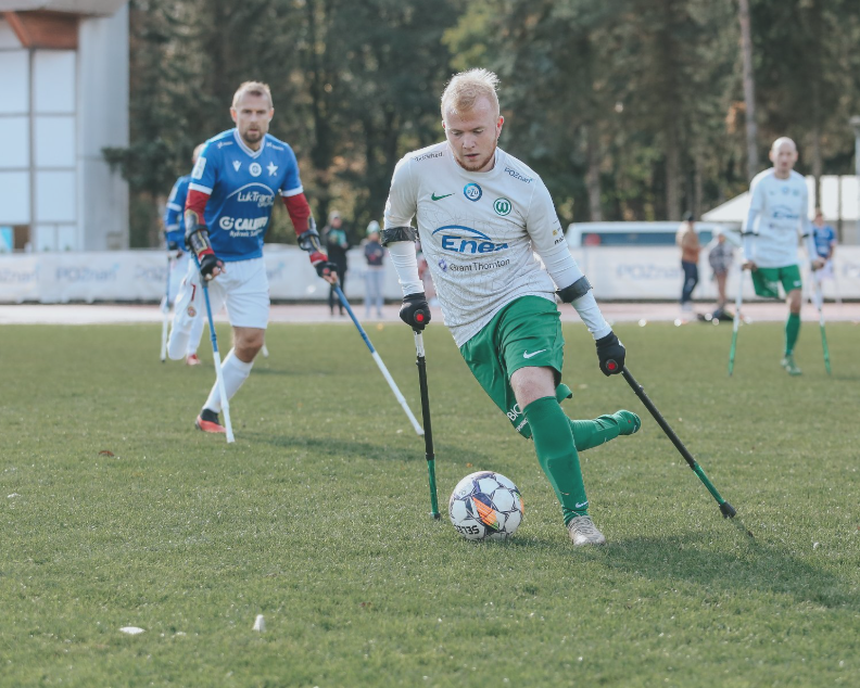 Zawodnicy sekcji amp futbolu Warty Poznań, fot. Tomasz Stefanik/Amp Futbol Polska Grupa sportowców gra w amp futbol na boisku trawiastym. Zawodnicy poruszają się, używając kul. Na pierwszym planie gracz w białej i zielonej koszulce z numerem 20 kontroluje piłkę lewą stopą. Ma na sobie zielone spodenki i zielone getry. W tle widoczny jest inny zawodnik w niebiesko-białym stroju oraz kilku graczy w biało-zielonych strojach. W tle drzewa i budynek.