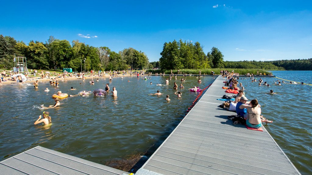 Zdjęcie przedstawia ludzi kąpiących się w jeziorze i siedzących na molo.