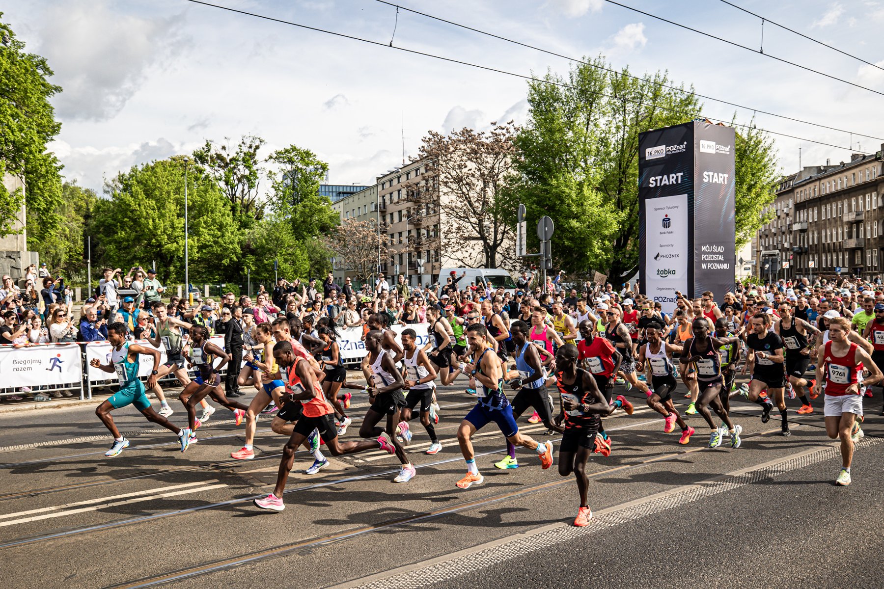 Coraz bliżej rekordu uczestników PKO Poznań Półmaratonu (fot. K. Berda) Maraton uliczny. Grupa biegaczy w połowie kroku uczestniczy w wydarzeniu sportowym. Po bokach drogi zgromadzeni widzowie kibicują i robią zdjęcia. W tle drzewa, budynki oraz duży baner z napisem ,START' i tekstem ,MÓJ ŚLAD POZOSTAŁ W POZNANIU'.