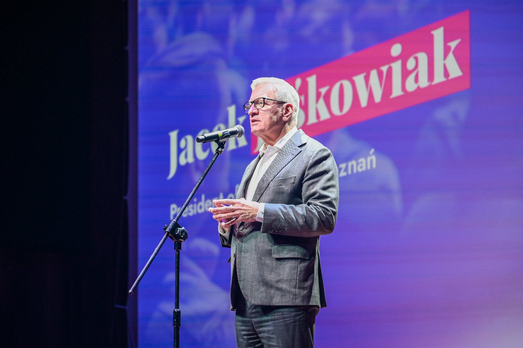 Mężczyzna w okularach i szarej marynarce stoi na scenie, przemawiając do mikrofonu. W tle fioletowy ekran z częściowo widocznym napisem: "Jacek Jaśkowiak" oraz "Prezydent Poznania".