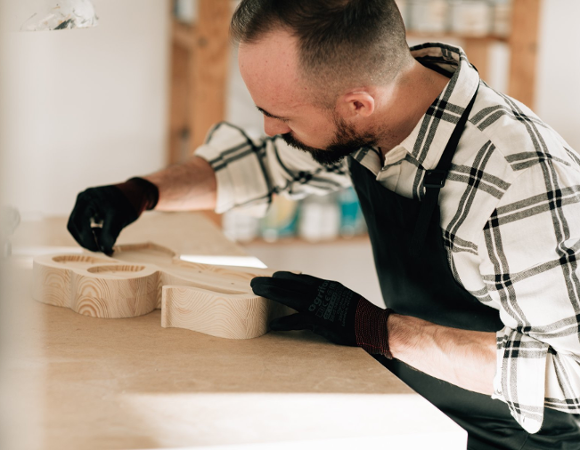 Mężczyzna z brodą, ubrany w koszulę w kratę i czarny fartuch ochronny, pracuje przy drewnianym stole warsztatowym. Ma założone czarne rękawice robocze i precyzyjnie obrabia dłońmi duży, jasny element wycięty z drewna o wyraźnym usłojeniu.