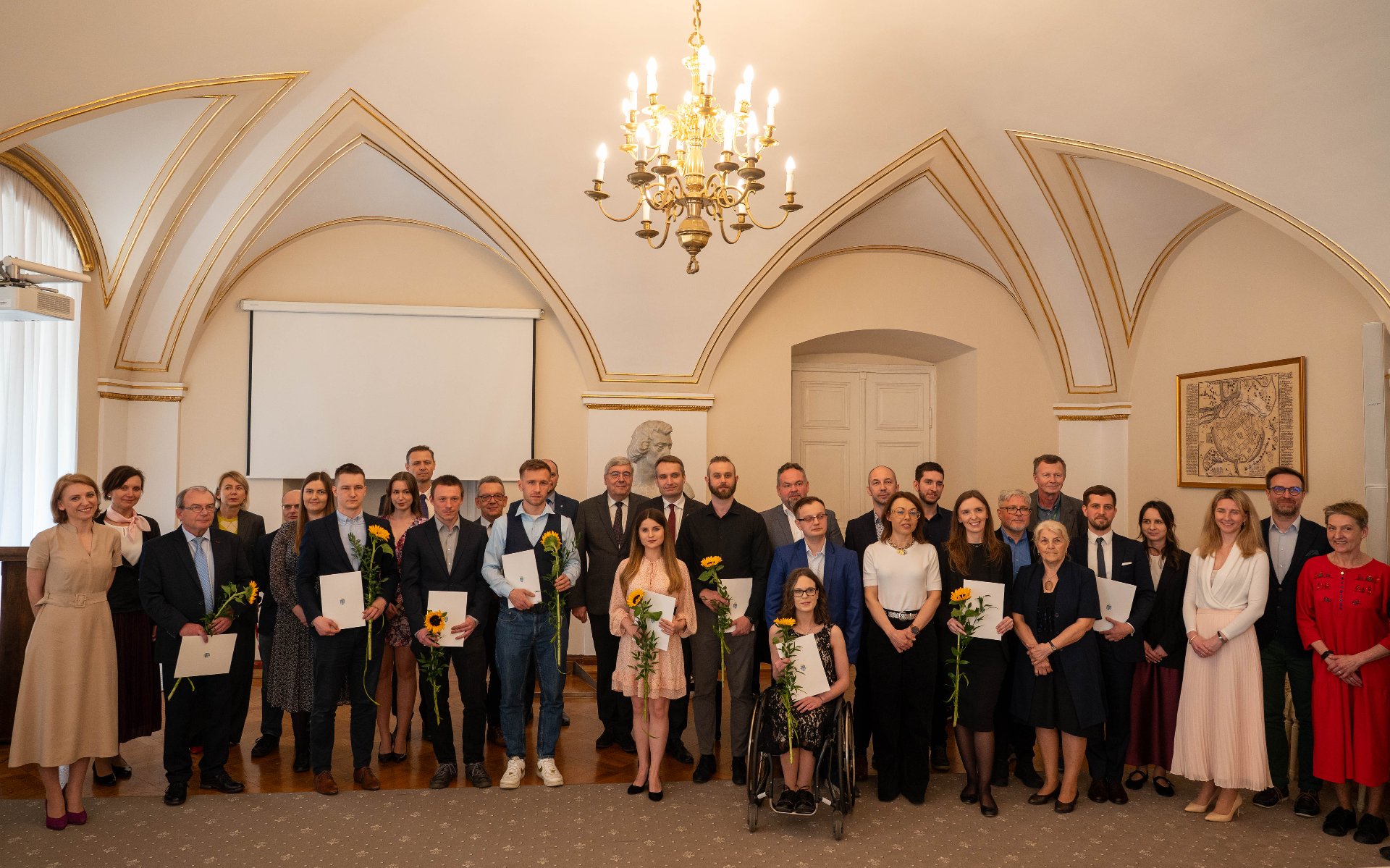 Już po raz dwudziesty pierwszy Miasto przyznało nagrody za najlepsze prace doktorskie i magisterskie Na białej kopercie leży słonecznik o jasnożółtych płatkach oraz zielona roślina o wydłużonych liściach.