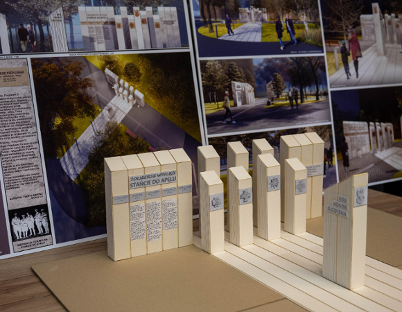 Na drewnianym stole leży plansza z wizualizacjami i tekstem, a przed nią stoi przestrzenna makieta pomnika. Makieta składa się z kilku beżowych prostopadłościanów symbolizujących kamienne tablice.
