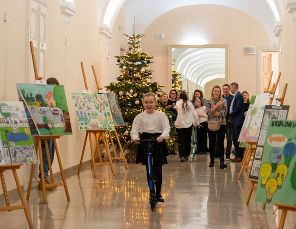 Na zdjęciu dziecko w korytarzu urzędu, jedzie na hulajnodze, obok widać prace plastyczne na sztalugach