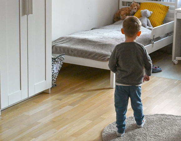 Na zdjęciu małe dziecko maszerujące w stronę łóżka, na którym leżą misie