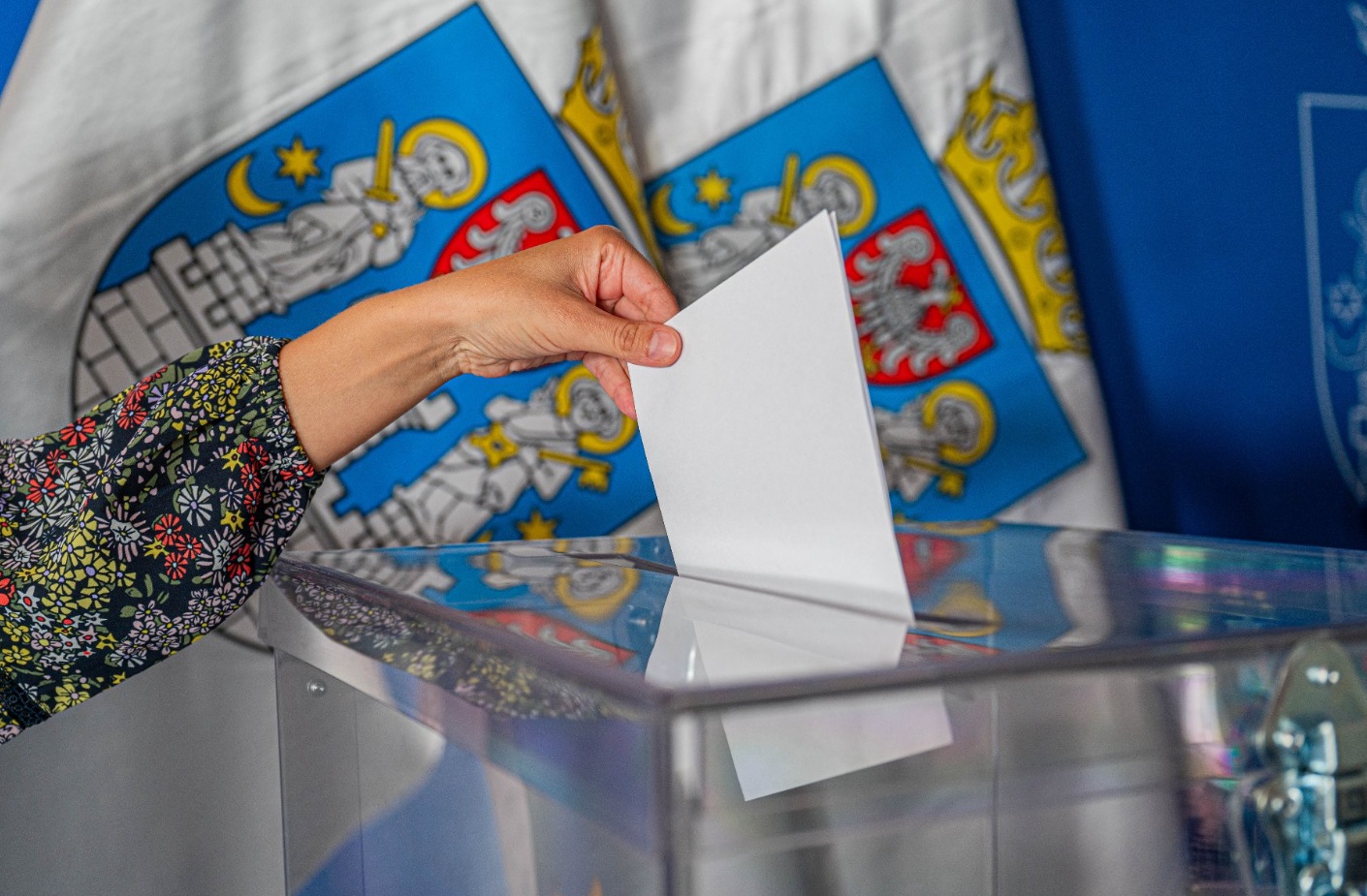 Już w najbliższą niedzielę odbędą się wybory do rad osiedli Na zdjęciu ręka wrzucająca głos do urny, w tle flagi z herbem Poznania