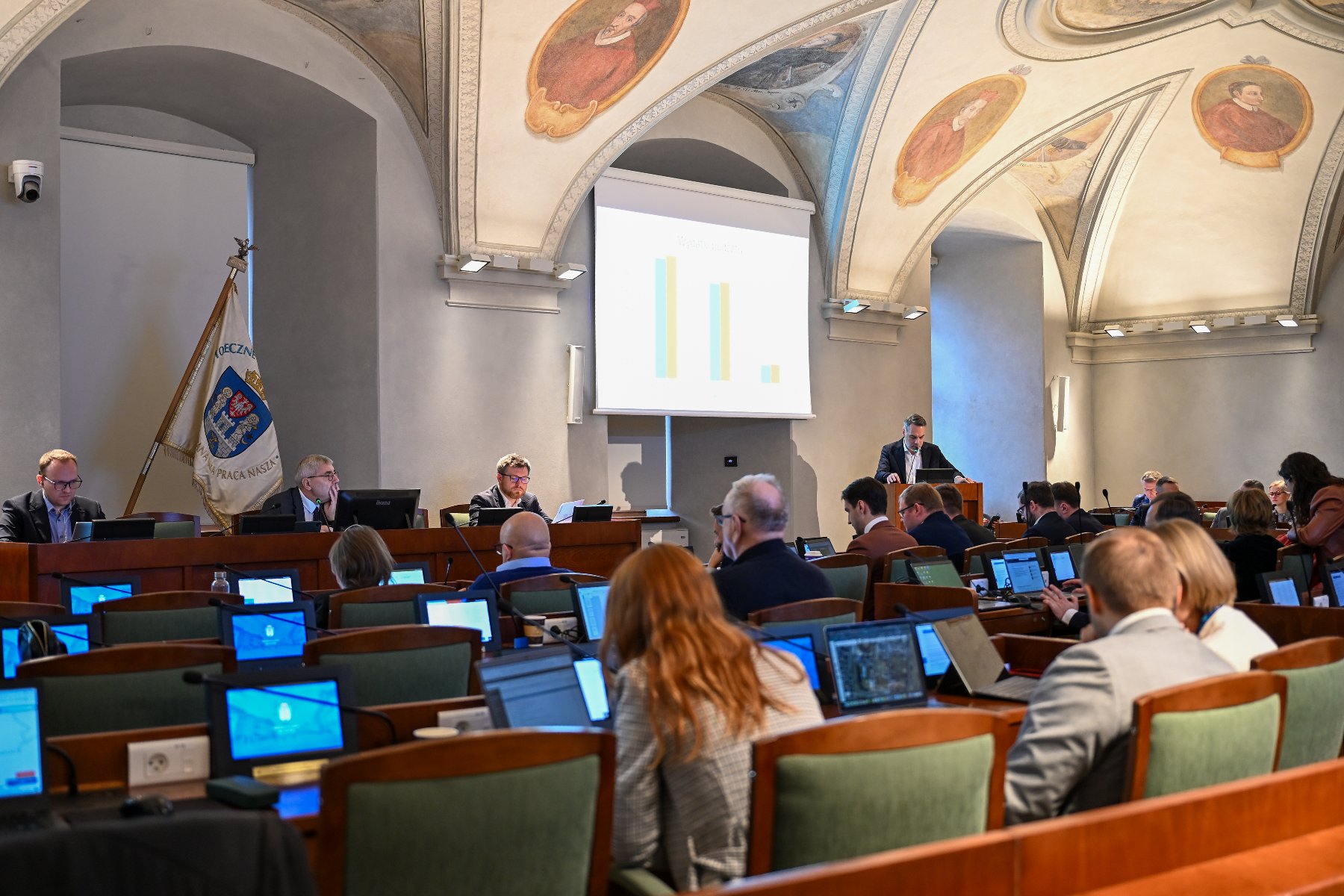 Na zdjęciu sala sesyjna, w niej siedzą radni, przy mównicy stoi mężczyzna