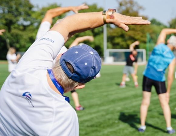 Na zdjęciu seniorzy uprawiający gimnastykę na świeżym powietrzu, stojący tyłem do obiektywu