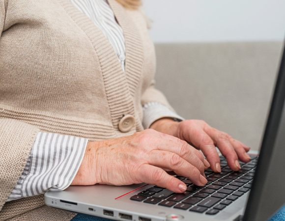 Na zdjęciu starsza osoba w swetrze pisząca coś na laptopie, widać tylko jej dłonie