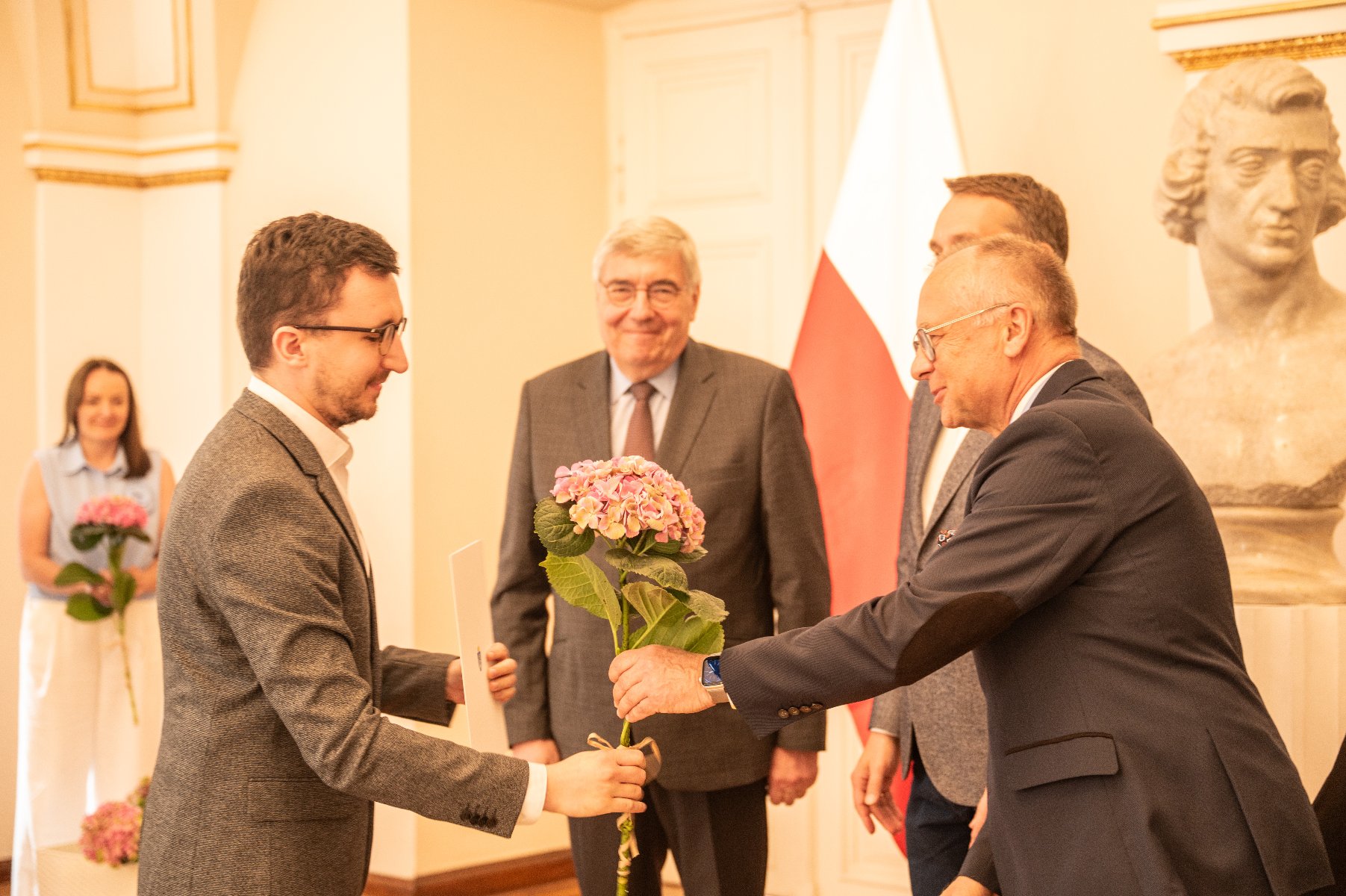 Stypendia naukowe Miasta Poznania przyznawane są od 1998 r. Na zdjęciu uroczystość wręczenia stypendiów. Trzech mężczyzn wręcza dyplom i kwiata.