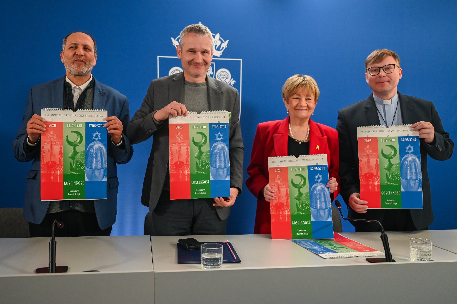Na zdjęciu widać cztery osoby stojące za jasnym stołem na tle niebieskiej ściany z białym godłem Poznania. Każda z osób trzyma przed sobą pionowy kalendarz zatytułowany "GOŚCINNOŚĆ. Kalendarz Trzech Religii".