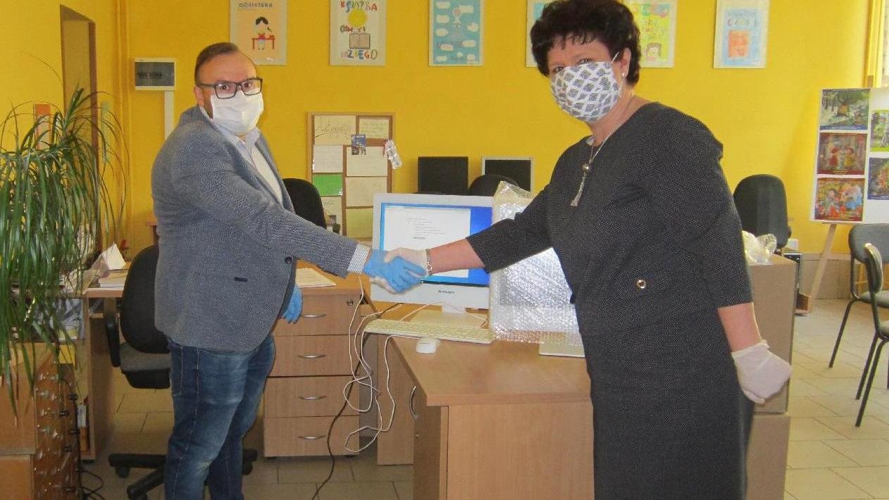 Na zdjęciu znajduje się darczyńca oraz dyrektorka szkoły, którzy podają sobie ręce. Na biurkach stoją komputery, na ścianie wiszą obrazki namalowane przez dzieci. - grafika artykułu