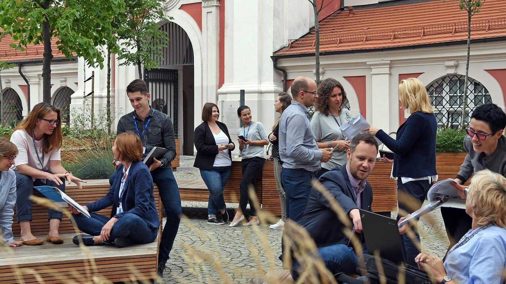Dziedziniec Urzędu Miasta Poznania. Na nim kilkanaście osób z identyfikatorami pracowników urzędu. Rozmawiają, mają w rękach teczki na dokumenty, pokazują sobie coś na laptopie.