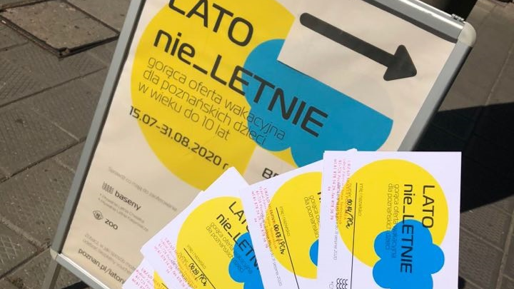 Na środku zdjęcia "potykacz" zapowiadający "LATO nie_LETNIE". Na pierwszym planie dłoń, trzymająca kilka voucherów na wydarzenia
