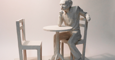 Projekt: Bohdan Smoleń siedzący na krześle przy niewielkim stoliku, zamyślony. Prawy łokieć opiera na stole i dłonią dotyka brody, lewa dłoń na oparciu krzesła. Naprzeciw drugie, puste krzesło