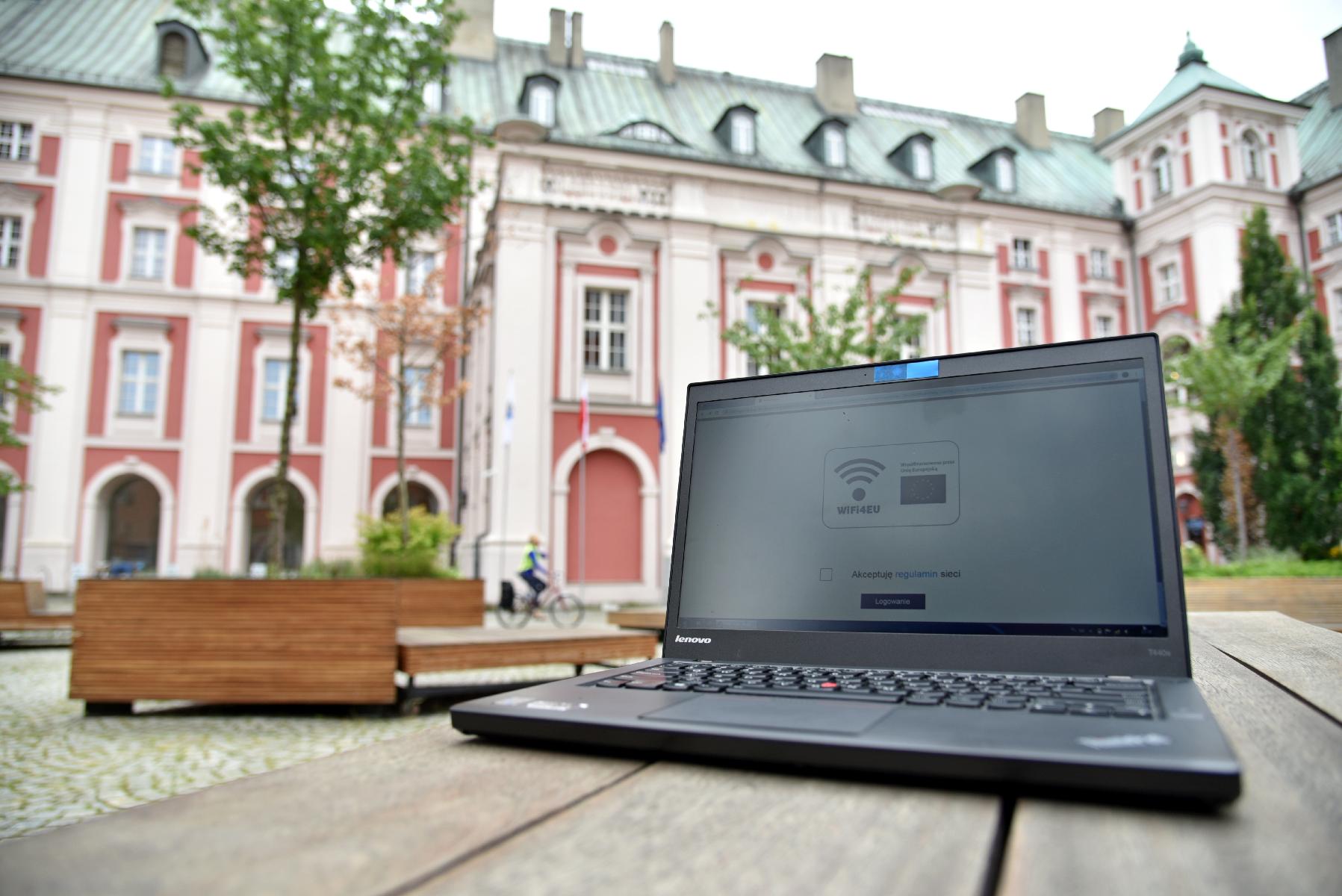 Zdjęcie przedstawia dziedziniec Urzędu Miasta Poznania. Na pierwszym planie widać komputer (laptop), stojący na ławce - na ekranie wyświetlona jest strona umożliwiająca połączenie z siecią, w tle widać budynek urzędu - grafika artykułu