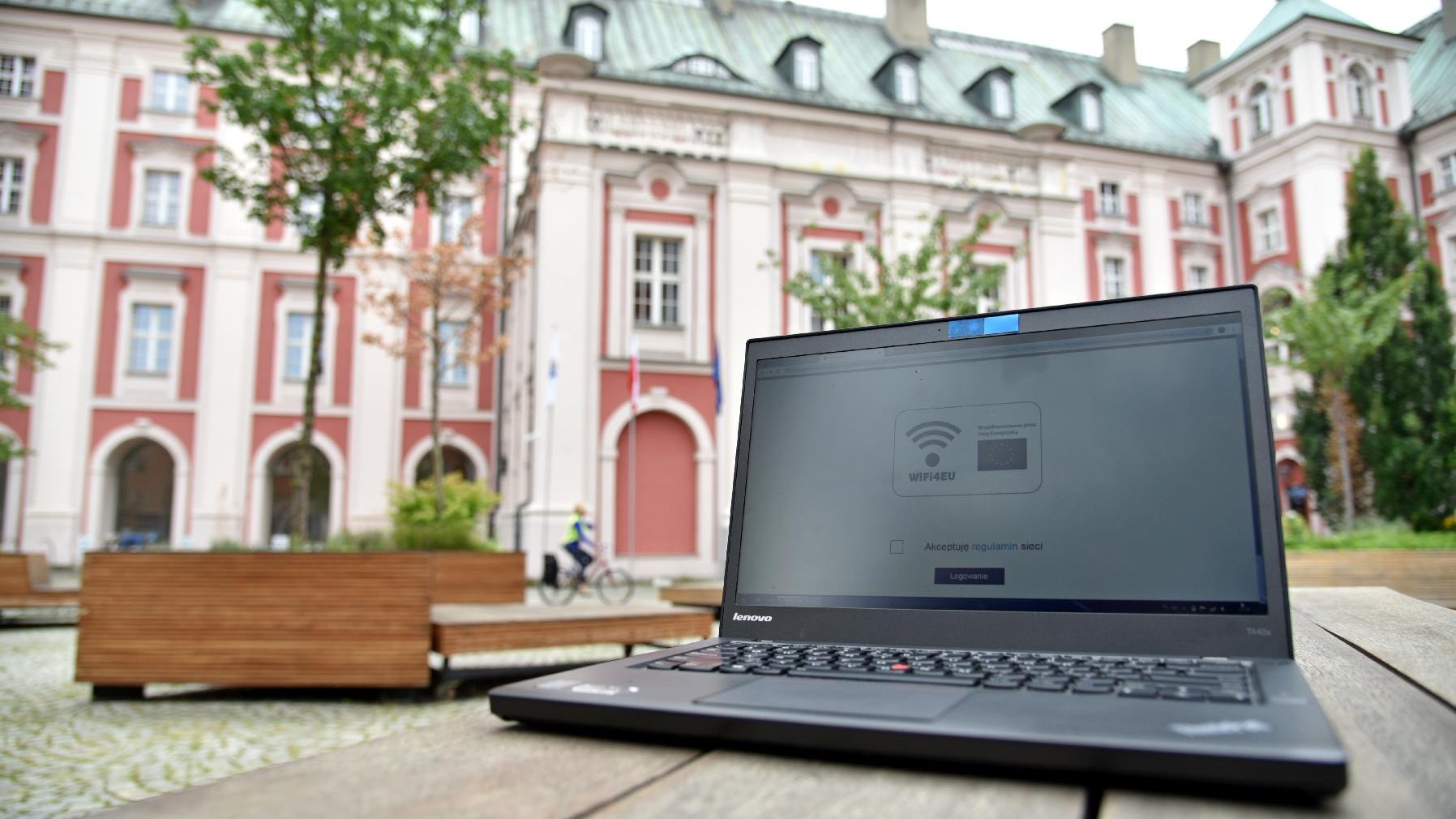 Zdjęcie przedstawia dziedziniec Urzędu Miasta Poznania. Na pierwszym planie widać komputer (laptop), stojący na ławce - na ekranie wyświetlona jest strona umożliwiająca połączenie z siecią, w tle widać budynek urzędu