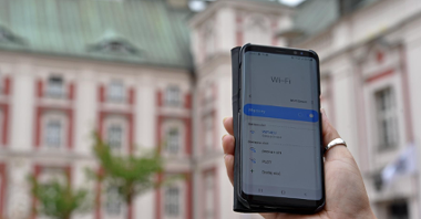 Zdjęcie przedstawia dziedziniec Urzędu Miasta Poznania. Na pierwszym planie widać trzymany w ręce telefon - na ekranie wyświetlona jest strona umożliwiająca połączenie z siecią, w tle widać budynek urzędu