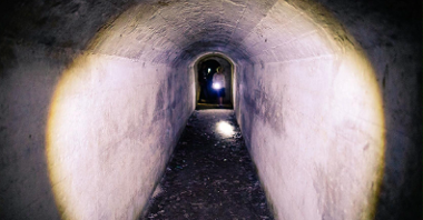 Na zdjęciu znajduje się wnętrze schronu, widać na nim tunel, a w oddali podążających nim ludzi