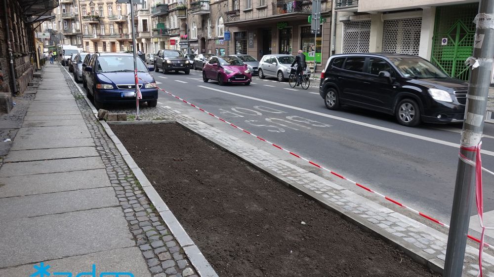 teren z ziemią przygptowany do nasadzeń. Po ulicy jada samochody i rowerzysta. Po obu stronach ulicy parkują równolegle samochody