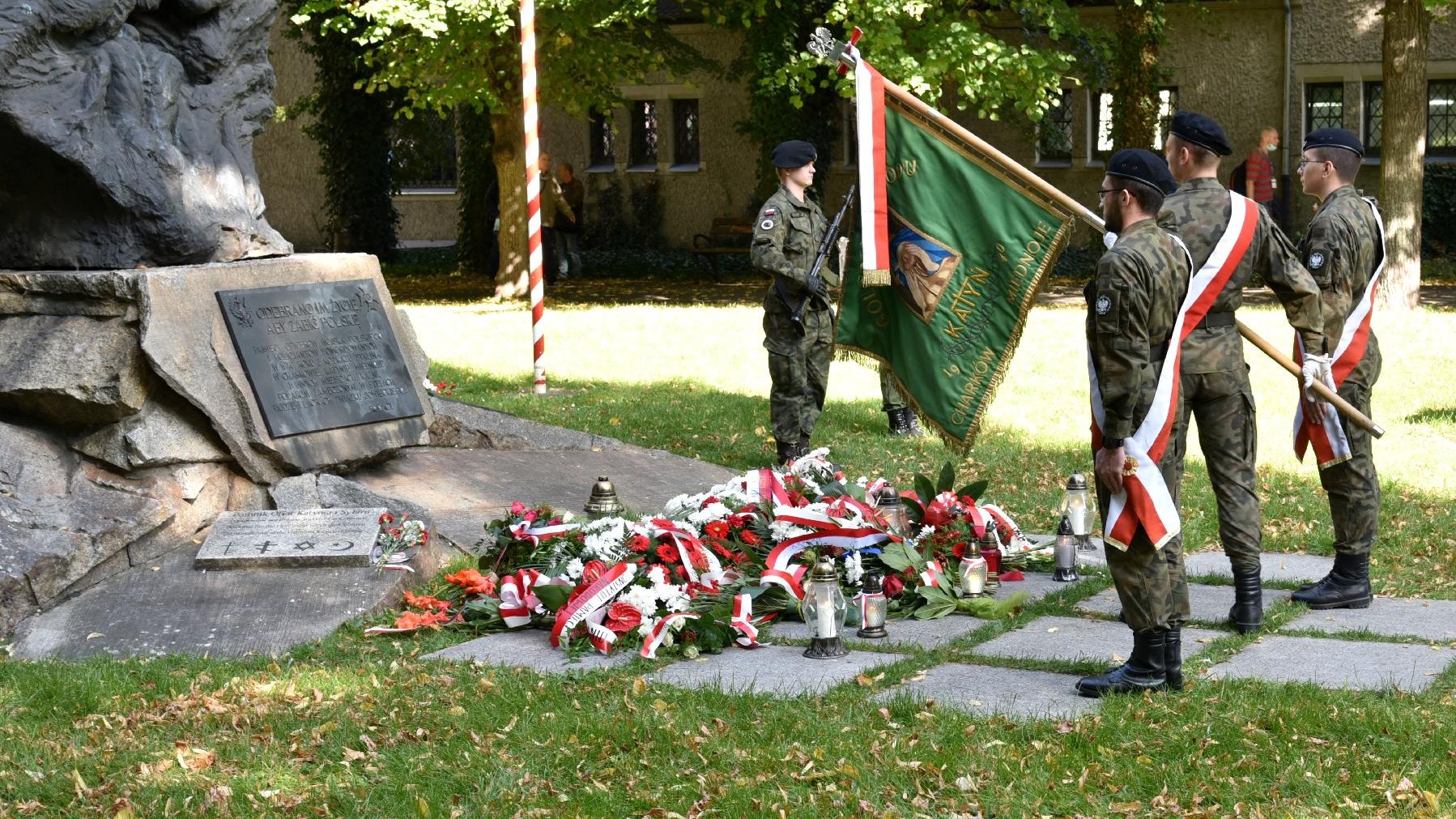 Zdjęcie przedstawia poczet sztandarowy i wiązanki kwiatów pod pomnikiem Ofiar Katynia i Sybiru.