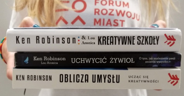 Kobieta trzymająca przed sobą trzy książki autorstwa Kena Robinsona - nie widać jej twarzy. Na bluzce ma napis: Forum Rozwoju Miast