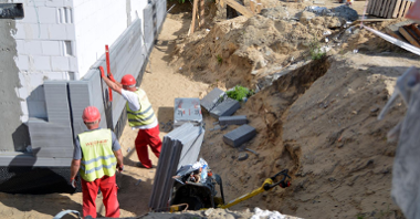 Wykop. W nim dwóch robotników w czerwonych kaskach przykłada narzędzia, mierząc warstwę muru budynku. W tle piach i materiały budowlane