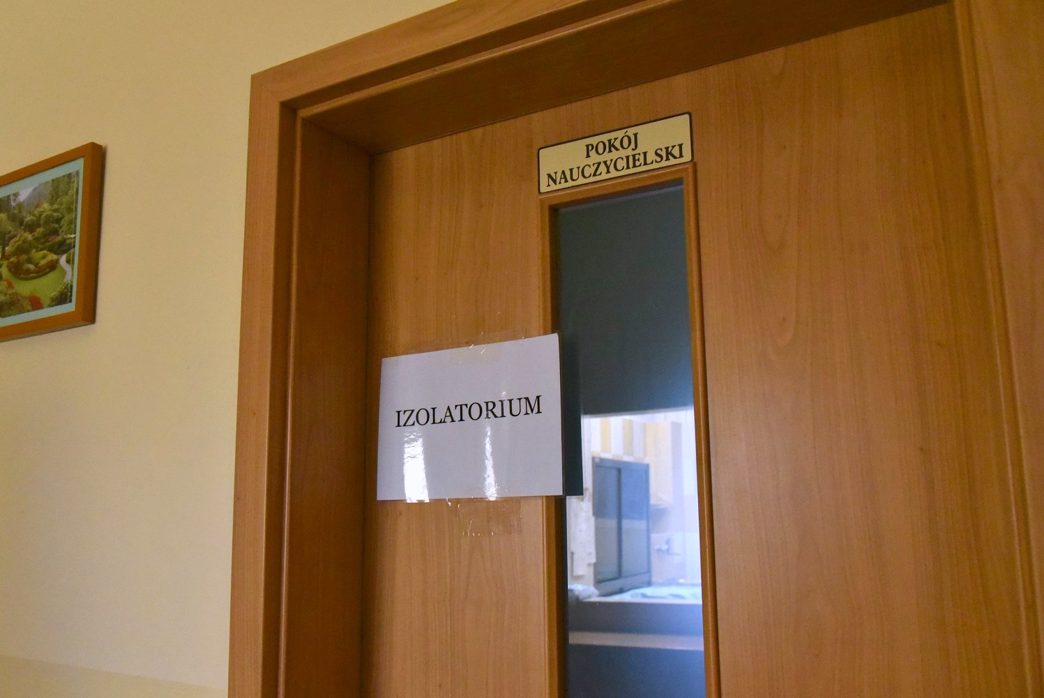 Zdjęcie przedstawia szkolne drzwi. Na nich tabliczka z napisem: Pokój nauczycielski. Poniżej przyczepiona laminowana kartka z napisem: Izolatorium - grafika artykułu