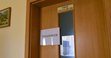 Zdjęcie przedstawia szkolne drzwi. Na nich tabliczka z napisem: Pokój nauczycielski. Poniżej przyczepiona laminowana kartka z napisem: Izolatorium