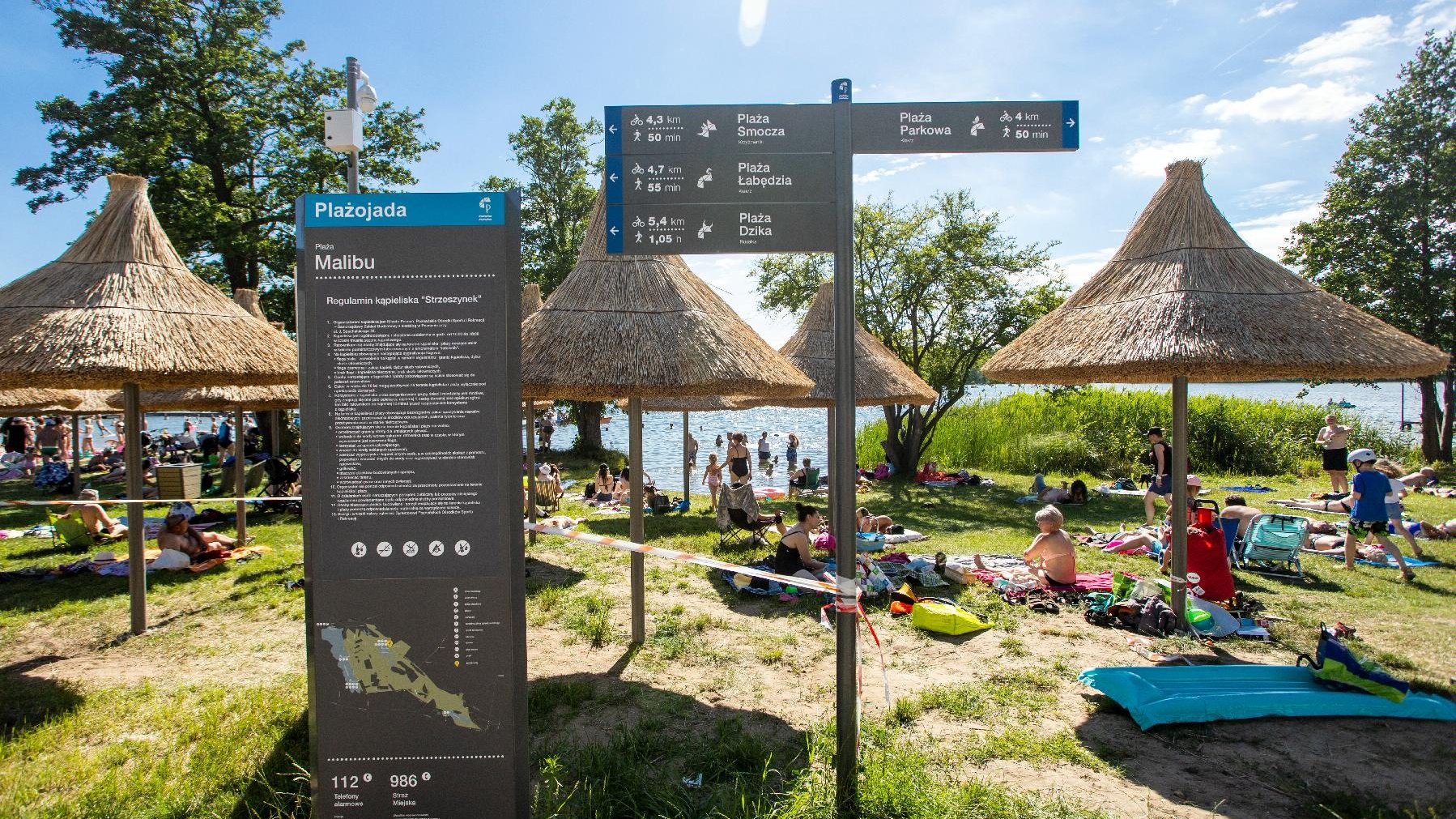 Zdjęcie przedstawia plażę - widać na nim siedzących na kocach ludzi, parasolki ze słomy oraz tablicę z opisem projektu Plażojada. - grafika artykułu