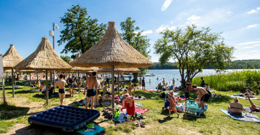 Zdjęcie przedstawia plażę - widać na nim siedzących na kocach ludzi, parasolki ze słomy oraz jezioro.
