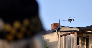 Zdjęcie przedstawia drona latającego nad budynkami. Na pierwszym planie widać głowę stojącego tyłem strażnika miejskiego.