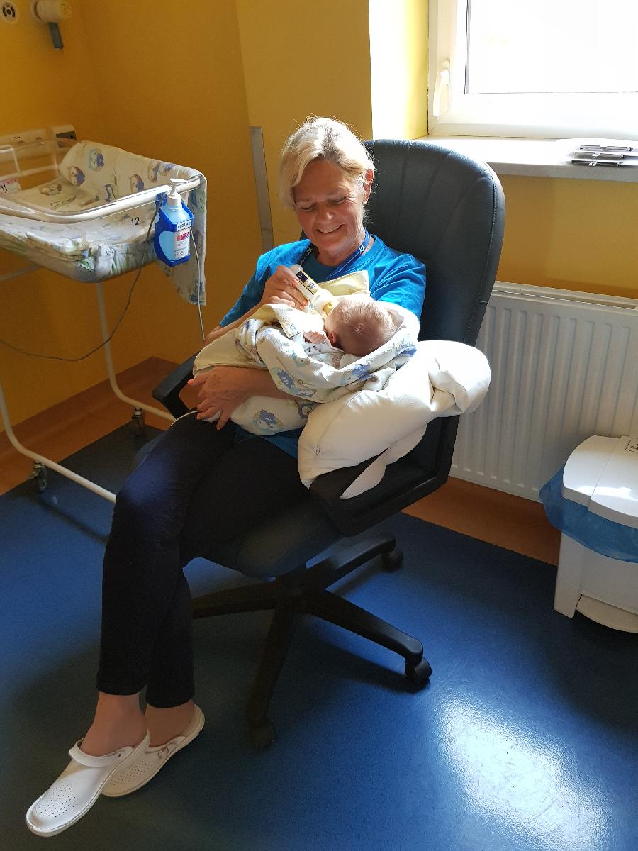 Wnętrze sali szpitalnej. Kobieta w wieku 60+ siedzi na krześle, uśmiechnięta. W ramionach trzyma noworodka, karmi go butelką - grafika artykułu