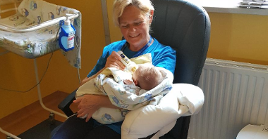 Wnętrze sali szpitalnej. Kobieta w wieku 60+ siedzi na krześle, uśmiechnięta. W ramionach trzyma noworodka, karmi go butelką