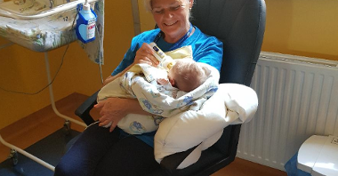 Wnętrze sali szpitalnej. Kobieta w wieku 60+ siedzi na krześle, uśmiechnięta. W ramionach trzyma noworodka, karmi go butelką