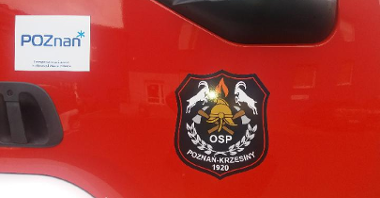 Czerwone dzwi do kabiny wozu strażackiego. Na nich naklejka "OSP Poznań-Krzesiny" oraz logo Miasta Poznania.