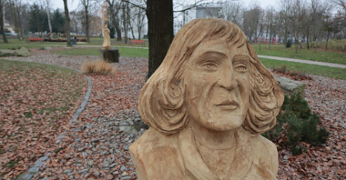 Zdjęcie przedstawia drewnianą rzeźbę Mikołaja Kopernika. W tle widać park oraz inną drewniana rzeźbę.