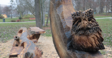 Zdjęcie przedstawia drewnianą rzeźbę sowy. W tle widać park.