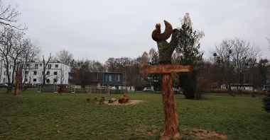 Zdjęcie przedstawia drewnianą rzeźbę z napisem "Park Heweliusza". W tle widać park.
