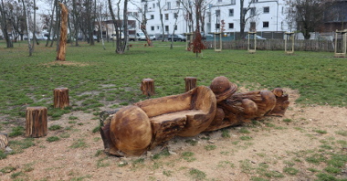 Zdjęcie przedstawia drewnianą rzeźbę pełniącą również funkcję ławki. W tle widać park.