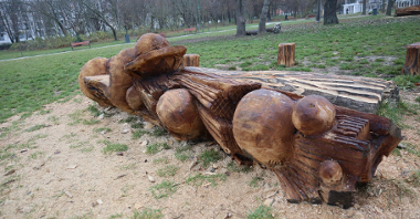 Zdjęcie przedstawia drewnianą rzeźbę pełniącą również funkcję ławki. W tle widać park.