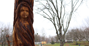 Zdjęcie przedstawia drewnianą rzeźbę dziewczyny. W tle widać park.