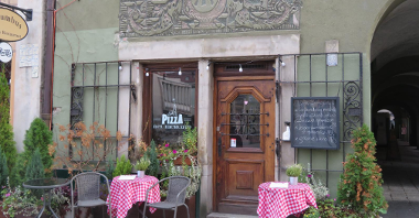 Zdjęcie przedstawia budynek pizzerii. Przed lokalem stoją stoliczki z czerwono - białym obrusem w kratkę i krzesła.