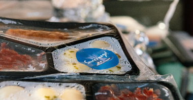 Zdjęcie przedstawia jedzenie w plastikowych opakowaniach. Na pudełkach naklejona jest naklejka z napisem "Smacznego".