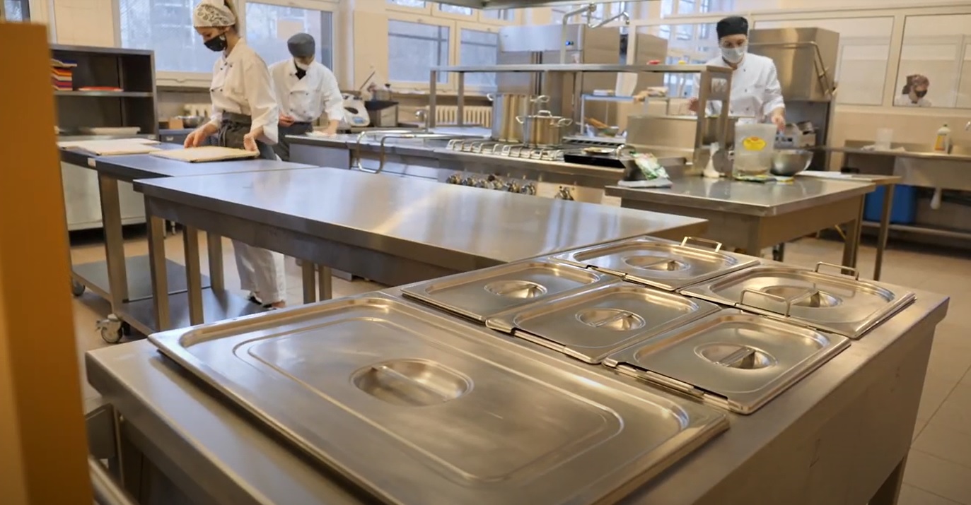 Szkolna pracownia wyglądająca jak kuchnia w restauracji. Na zdjęciu widać metalowe stoły, przy których pracuje trójka uczniów. - grafika artykułu